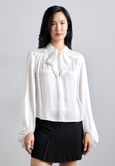 Blusa blanca con un escote de lazo, mangas largas abullonadas y hombros fruncidos. Combinada con una falda negra con pliegues. Tela suave y ligera.