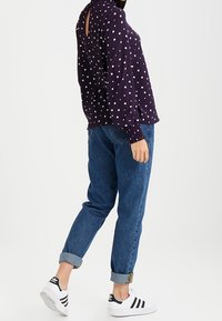 Personne portant un jean bleu retroussé, des baskets blanches avec des rayures noires et un haut à manches longues violet avec des pois blancs.