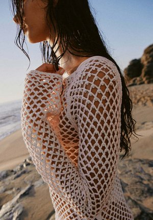Mujer con el pelo mojado que lleva un top blanco de crochet de manga larga, de pie en una playa de arena cerca de rocas al atardecer.