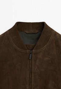 Bruine suède bomberjack met een geribde kraag en ritssluiting. Gladde textuur en minimale details. Voering aan de binnenkant van donker stof.