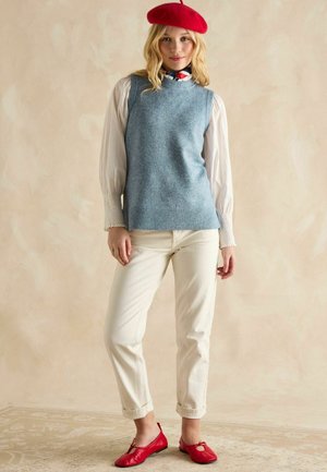 Hellblau gestrickter ärmelloser Pullover über einer weißen Bluse mit Puffärmeln, kombiniert mit cremefarbenen Hosen und roten flachen Schuhen. Das Modell trägt eine rote Baskenmütze.