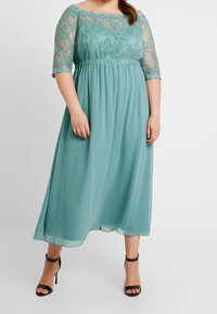 En lång, teal klänning med spetsärmar och figursydd midja. Kjolen är lätt och flowy, gjord av ett lättviktig tyg, och har en scalloped spetsdesign.