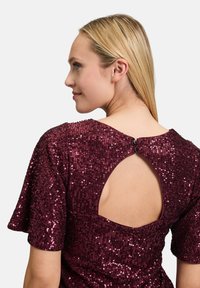 Abito in sequin borgogna con maniche corte, schiena a chiave con chiusura a bottone e tessuto texturizzato che riflette la luce.