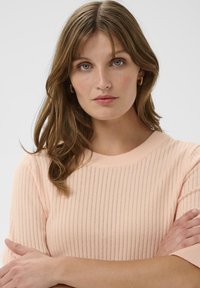 Strickpullover in Pfirsich mit horizontalen Rippstrukturen, rundem Halsausschnitt und kurzen Ärmeln. Das Modell hat die Arme verschränkt und zeigt die Textur des Kleidungsstücks.