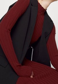 Personne portant une robe col roulé maroon côtelée superposée avec un blazer sans manches noir, assise les mains jointes devant.
