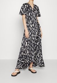 Robe longue noire avec motif abstrait en coups de pinceau blancs, manches courtes évasées, encolure en V et ourlet volanté. Portée avec des sandales plates.