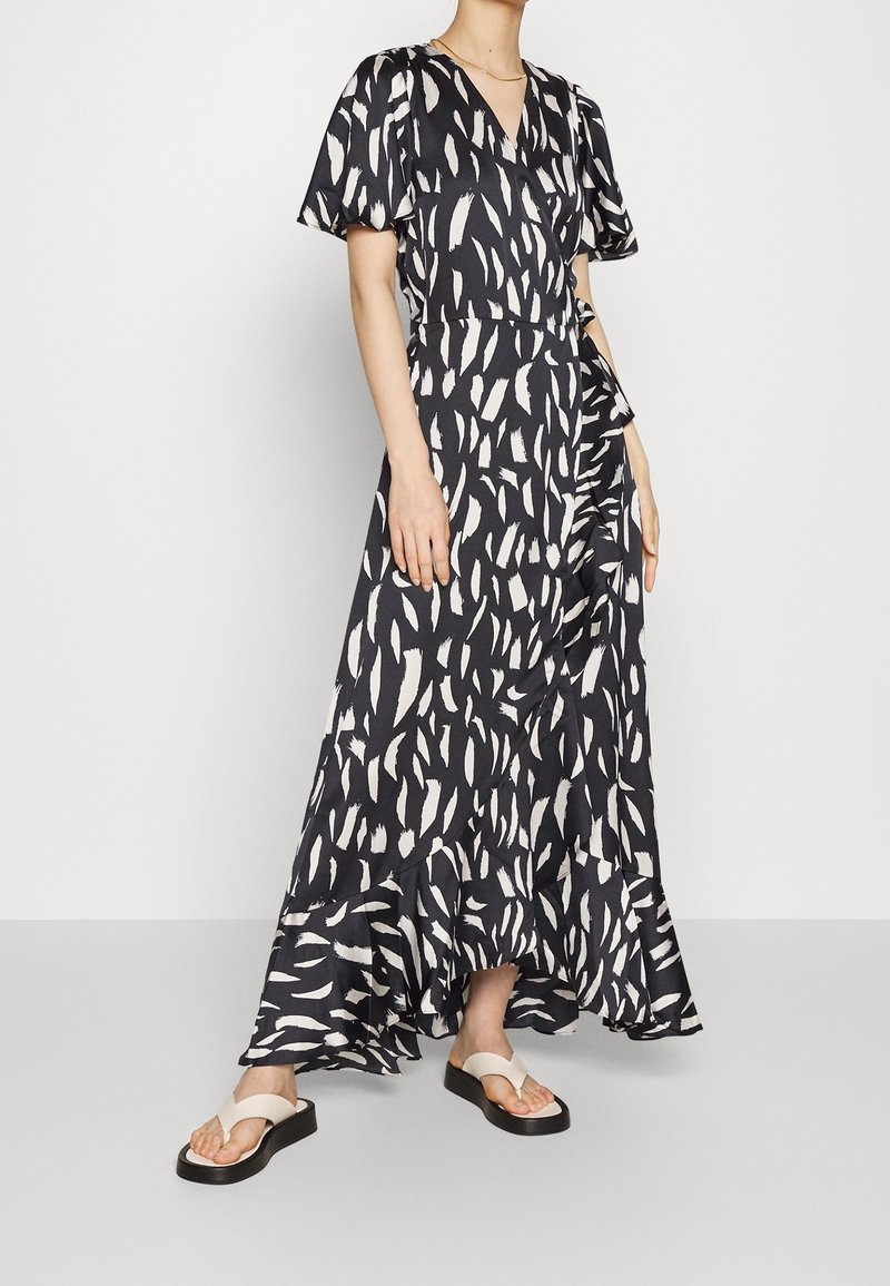 Robe longue noire avec motif abstrait en coups de pinceau blancs, manches courtes évasées, encolure en V et ourlet volanté. Portée avec des sandales plates.