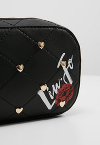 Musta tikattu keinonahkainen kosmetiikkalaukku, jossa on kultaiset sydännastat ja valkoinen teksti. Sisältää punaisen suudelmajäljen ja pyöristetyt reunat.