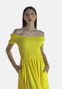 Gul off-shoulder kjole med smocket skørt, korte puffærmer og flydende silhuet. Har sidelommer og letvægtsstof.