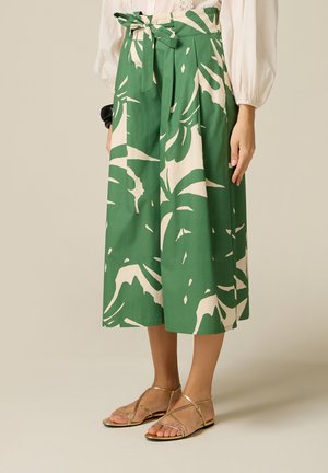 Femme portant un pantalon large imprimé de feuilles vertes et blanches avec taille nouée, un chemisier blanc à manches bouffantes et des sandales dorées à brides.