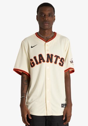 Uomo in piedi che indossa una maglia da baseball color crema con la scritta "GIANTS" in lettere nere e arancioni e pantaloni neri su uno sfondo bianco semplice.