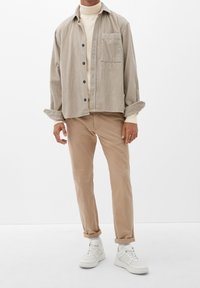 Chemise en velours côtelé beige clair avec boutons noirs, associée à un col roulé crème et un pantalon beige, complétée par des baskets blanches.