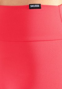 Rode sportlegging gemaakt van glad, rekbaar materiaal met een brede tailleband en een zwarte merklabel bovenaan.