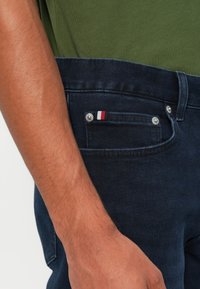 Tmavě modré džíny s přední kapsou, která má stříbrné kovové detaily a malý pruhovaný prvek v červené, bílé a modré barvě. Zelená košile je vidět nahoře.