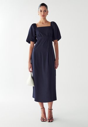 Robe bleu marine de longueur mi-longue avec des manches bouffantes, décolleté carré et coupe ajustée ; le modèle tient un petit sac à main blanc et porte des talons noirs à lanières.