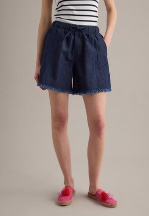 Donna con pantaloncini di jeans blu scuro con orli sfilacciati, top senza maniche a righe e scarpe slip-on rosa con nappe.