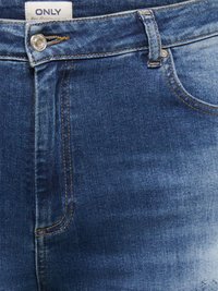 Gros plan sur un jean en denim bleu montrant la poche avant, la passants de ceinture, un bouton métallique avec le tampon de la marque et une étiquette blanche avec le texte "ONLY".