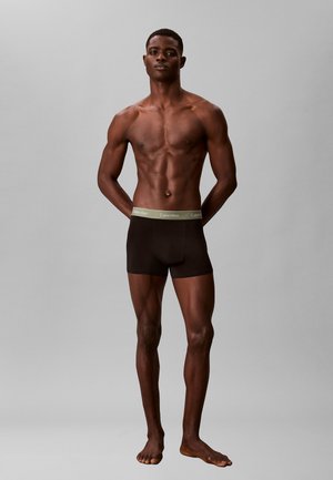 Uomo dalla pelle scura, in piedi scalzo con le mani dietro la schiena, indossa boxer neri Calvin Klein su uno sfondo grigio uniforme.