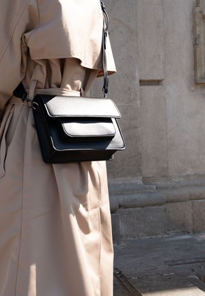 Persoon draagt een beige trenchcoat en draagt buiten bij een stenen muur een zwarte leren crossbodytas.