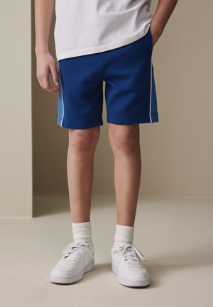 Isik, kes kannab siniseid sportluspükse valgete küljeribadega, valgeid keskmise pikkusega sokke ja valgeid tossusid, seisab toas beežise seina ääres.