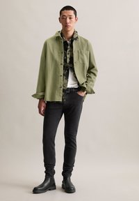 Green textured overshirt over a black camo shirt, paired with black skinny jeans and black chelsea boots. Model stands against a neutral background.