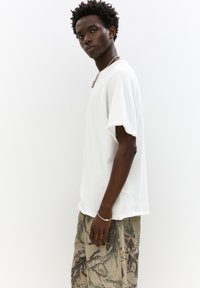 Hvid bomulds t-shirt med korte ærmer, parret med mønstrede grønne og beige shorts med naturinspirerede designs. Modellen står sidelæns.