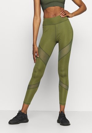 Leggings vert olive avec découpes en maille et coutures contrastées, taille haute, tissu extensible, associés à des chaussures de sport noires.