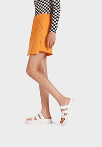 Mini gonna in corduroy arancione con orlo sfilacciato, abbinata a sandali in gomma bianchi con ritagli e cinturini regolabili. Top a scacchi in bianco e nero.