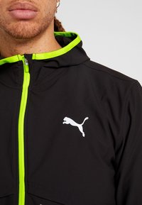 Gros plan d'une personne portant une veste noire avec une fermeture éclair vert néon et une doublure de capuche, avec un logo Puma blanc sur la poitrine.