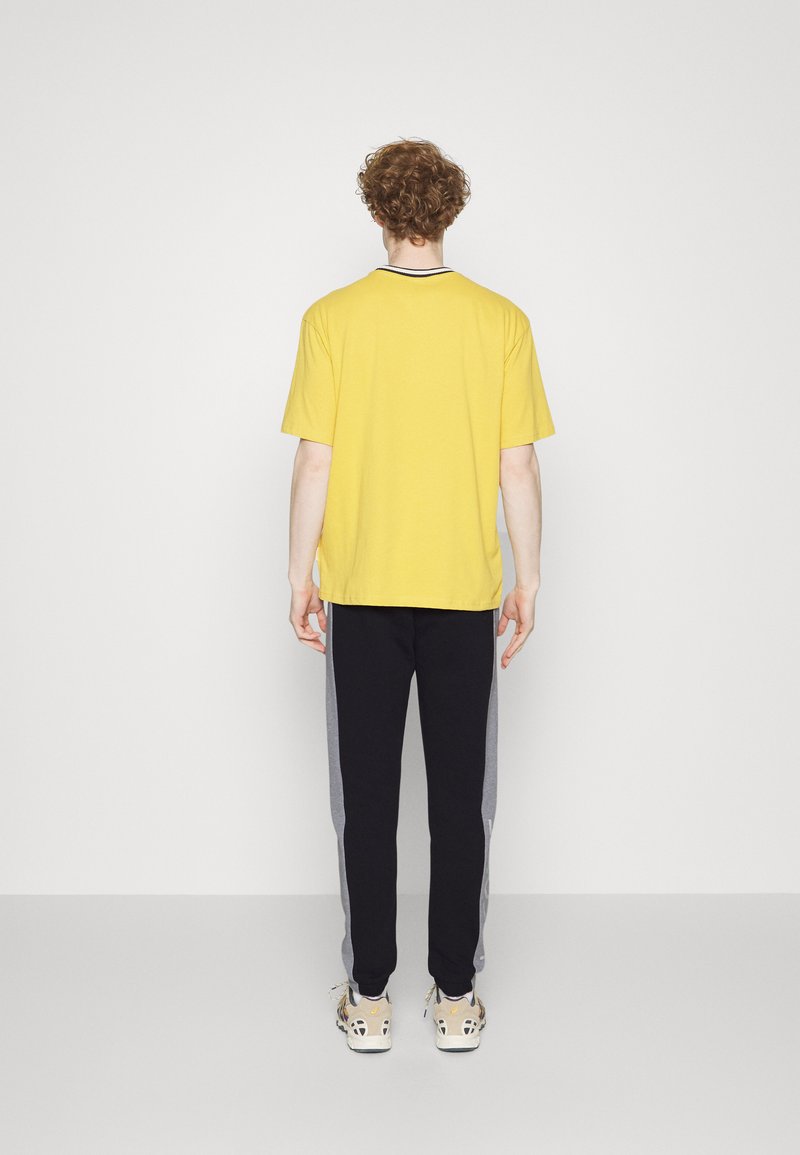 T-shirt en coton jaune, à manches courtes, coupe décontractée ; associé à un pantalon de jogging noir avec des panneaux latéraux gris et des baskets de couleur claire.