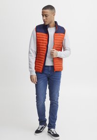 Polstret vest i orange med navy overdel, fuld lynlås, ståkrave og let tekstur. Sammen med grå sweater og blå jeans.