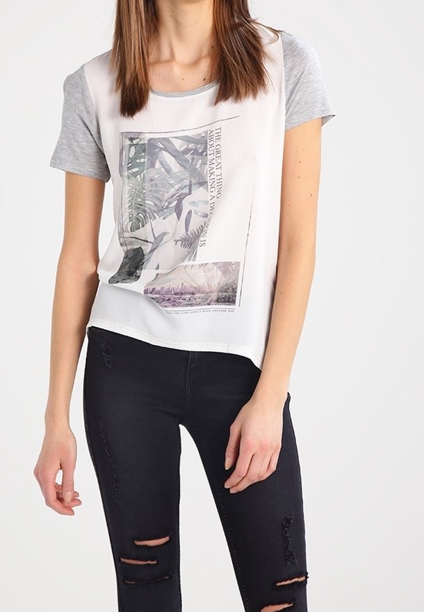 Femme portant un t-shirt graphique blanc et gris à manches courtes et un jean skinny noir déchiré, debout devant un fond uni.