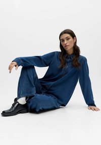 Femme aux longs cheveux bruns assise par terre, portant une chemise en denim bleu foncé à boutons et un pantalon assorti brodé, avec des chaussures noires.