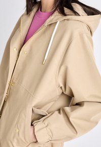 Beżowa kurtka z kapturem z złotymi akcentami, dwoma bocznymi kieszeniami, elastycznymi mankietami i sznurkiem; noszona na różowym topie. Gładka faktura.