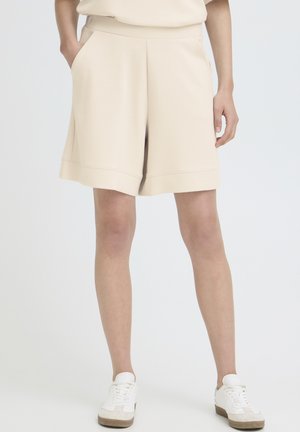 Ember bézs színű, térdig érő, zsebes rövidnadrágot és fehér sportcipőt visel, áll, lazán oldalra engedett kézzel.