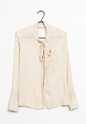 Kremowa bluzka z długim rękawem, z guzikami z przodu i wiązaniem w kokardę przy dekolcie, wisząca na czarnym wieszaku na białym tle.