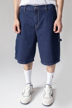 Blue denim shorts with a loose fit, featuring front and side pockets, visible stitching, and a mid-thigh length. Paired with white socks and sneakers.