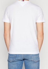 Bílý bavlněný tričko, krátké rukávy, kulatý výstřih, obsahuje malý trojúhelníkový akcent na zádech se červenými, bílými a modrými pruhy.