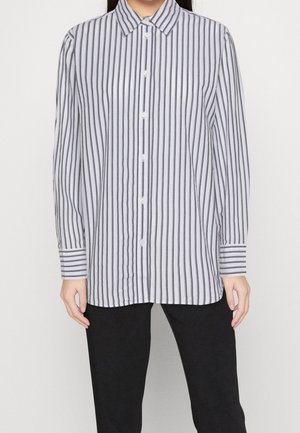 Femme portant une chemise boutonnée blanche et noire à rayures verticales avec manches longues et un pantalon noir, debout devant un fond blanc.