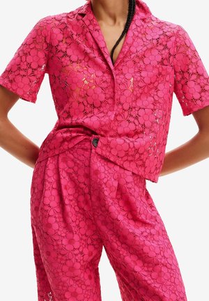 Helles pinkes Spitzen-Shortsleeve-Shirt und passende hoch taillierte Hosen mit Blumenmuster, getragen von einer Person mit Händen in der Hüfte.