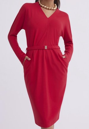 Rotes langärmliges Kleid mit V-Ausschnitt, tailliertem Gürtel, Taschen und glatter Textur. Auffällige goldene Halskette sorgt für einen kontrastierenden Akzent.