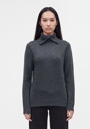 Kvinde med langt mørkt hår iført en mørkegrå sweater med høj krave og sorte bukser, stående foran en ensfarvet lys baggrund.