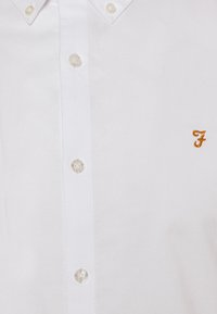 Bílá košile na knoflíky s malým oranžovým vyšívaným písmenem "F" na prsou, s límečkem zapínaným na knoflíky.