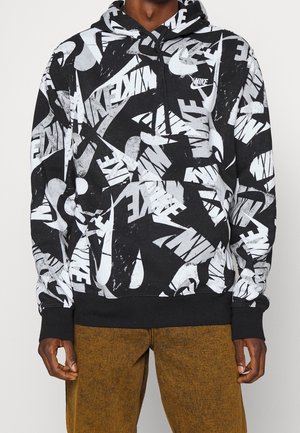 Mies, joka pukeutunut mustavalkoiseen Nike-huppariin, jossa on näkyvä logopainatus, ja ruskeisiin ribbineuloshousuihin, seisoo yksinkertaisen taustan edessä.