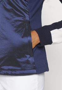 Veste bleu foncé avec un panneau de poche brillant, main glissée à l'intérieur, portée sur un haut à manches longues blanc et marine, associée à un pantalon blanc.