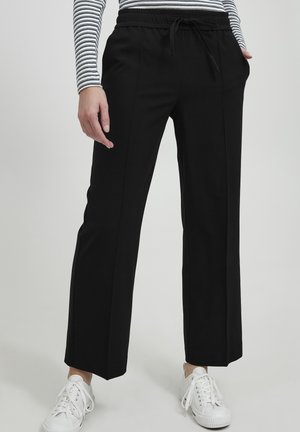 Black, wide-leg trousers with an elastic waistband and drawstring, featuring two side pockets and a smooth texture. Worn with white sneakers.
