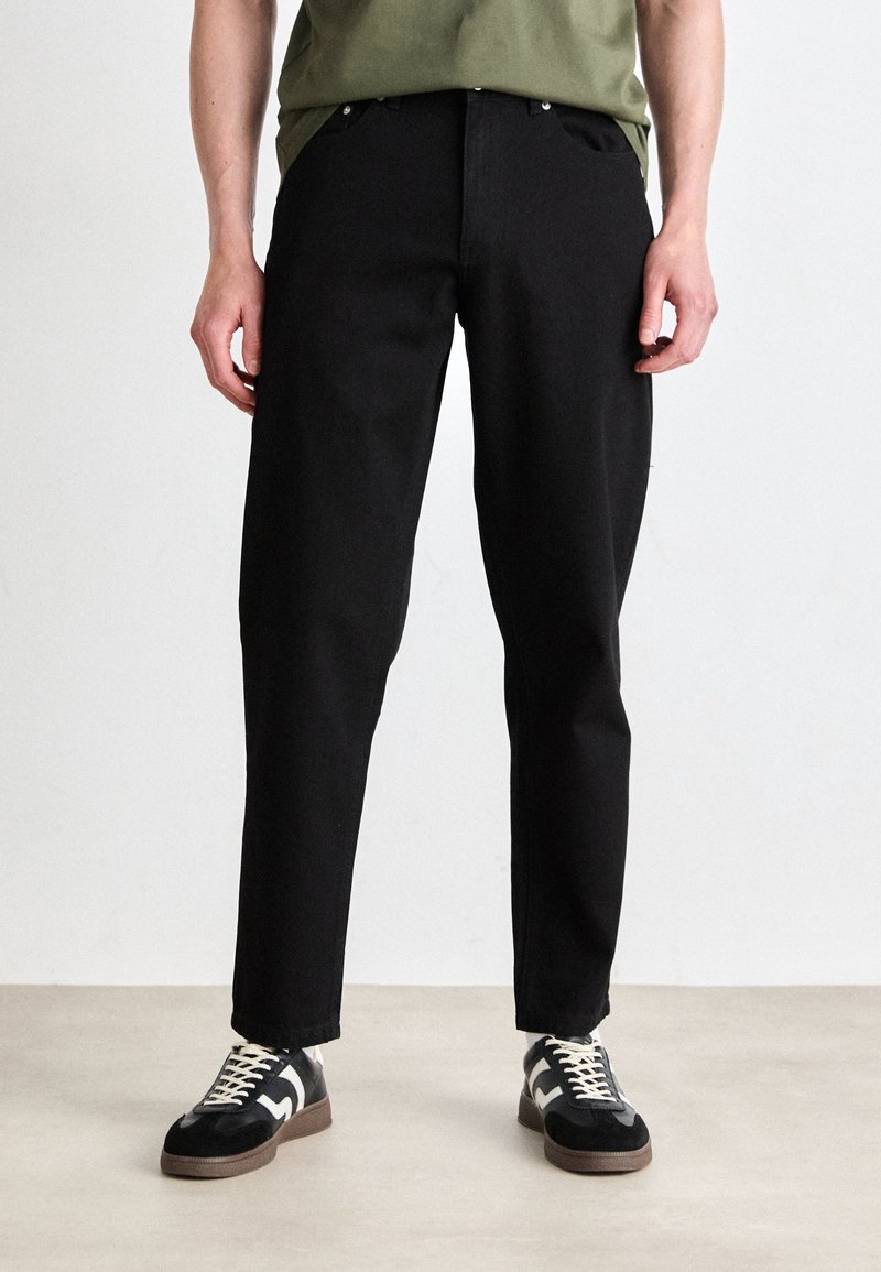 Homme portant un pantalon noir droit, une chemise verte et des baskets noires à lacets blancs, debout sur un sol beige devant un mur uni.