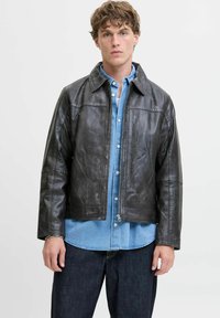 Chaqueta de cuero negra con corte recto y cierre de cremallera, que presenta un cuello con solapa y detalles de costura horizontal. Llevada sobre una camisa de denim azul claro.