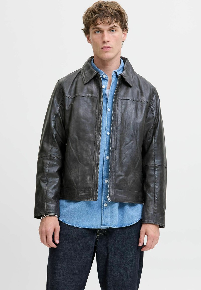Chaqueta de cuero negra con corte recto y cierre de cremallera, que presenta un cuello con solapa y detalles de costura horizontal. Llevada sobre una camisa de denim azul claro.