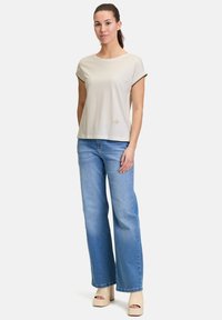 Lys beige bomuld t-shirt med rullede ærmer, en rund halsudskæring og en lille broderet detalje. Matchet med vide blå denim jeans.